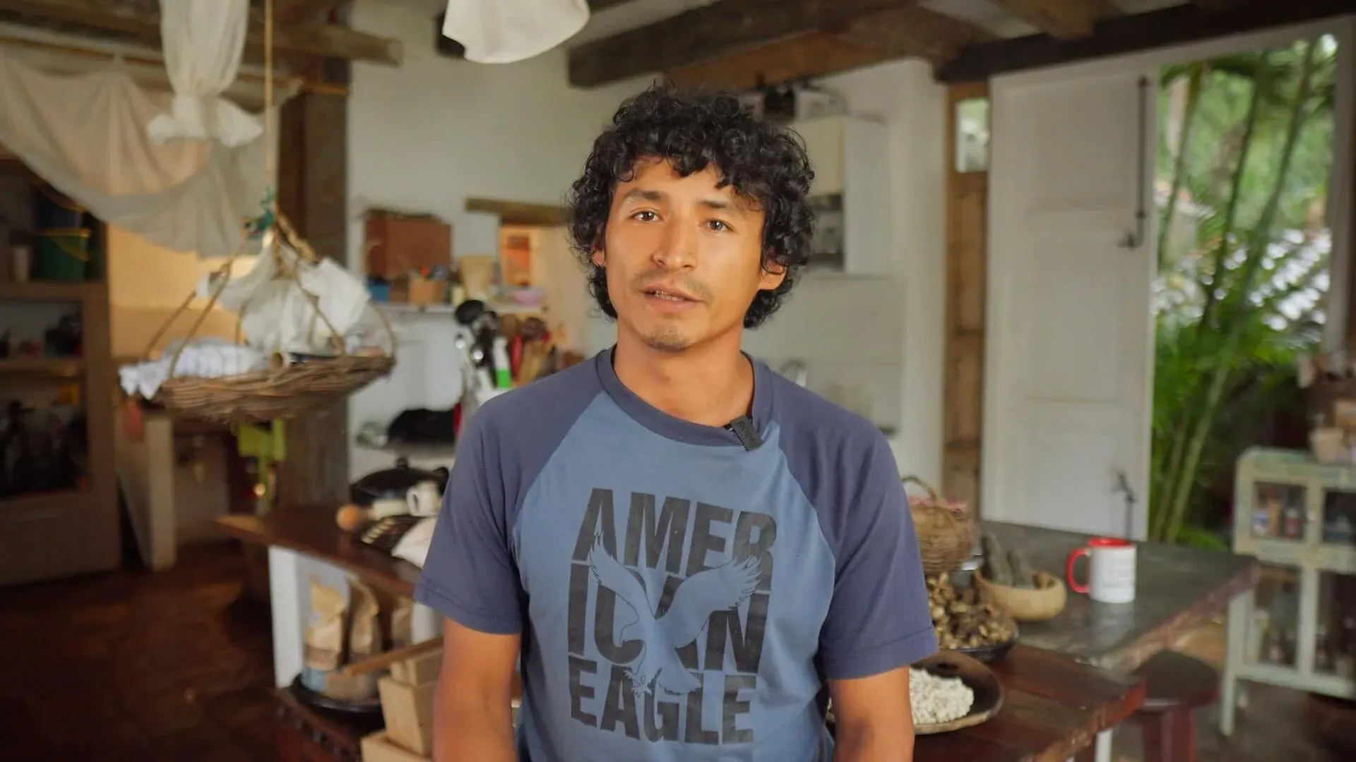 Man in Casa Común, wearing an American Eagle shirt, standing in a rustic kitchen.