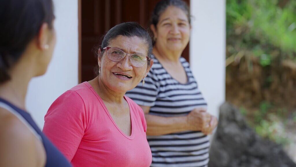 Three women talking outside a house. One wears a pink shirt, another a striped shirt.
