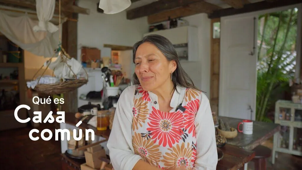Casa Común: A woman in a floral apron smiles in a rustic kitchen.
