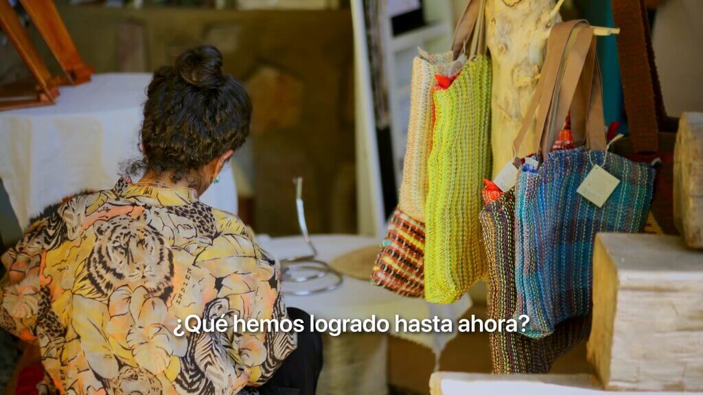 Artisan with colorful woven bags. Text: "¿Qué hemos logrado hasta ahora?