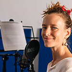A person with a red ribbon in their hair smiles while standing in front of a microphone and music stand, showcasing their voice talent in the recording studio.