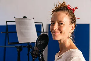 A person with a red ribbon in their hair smiles while standing in front of a microphone and music stand, showcasing their voice talent in the recording studio.