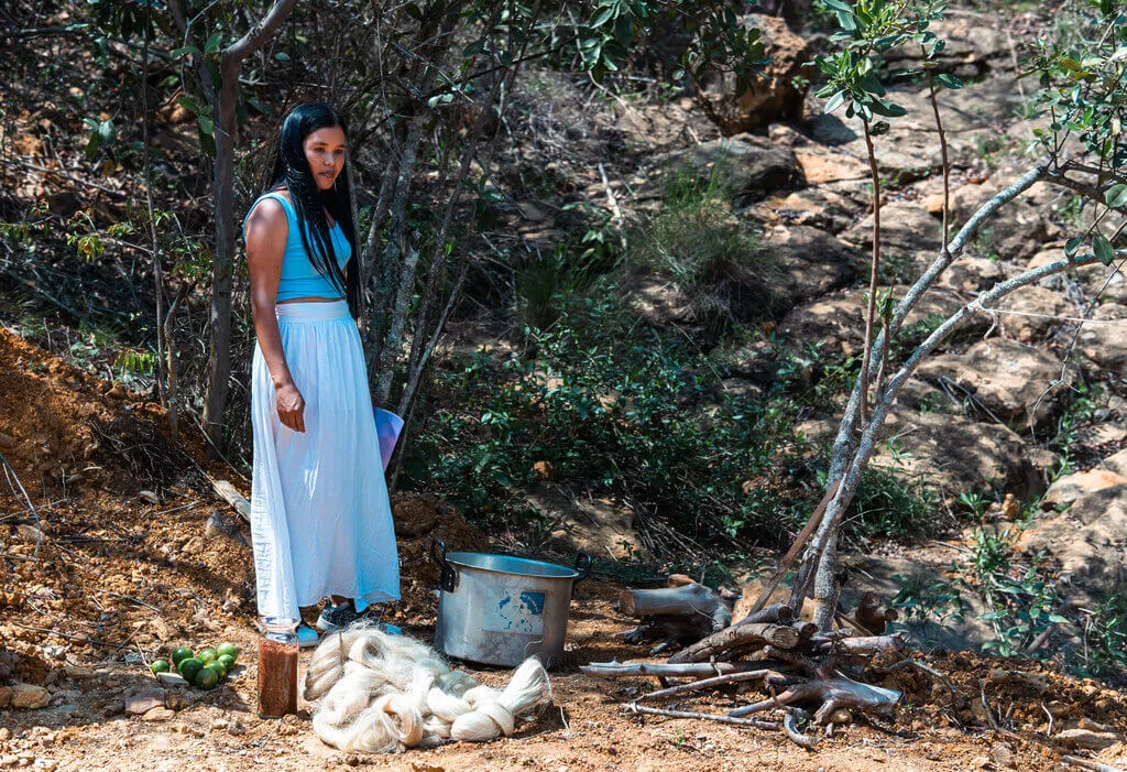 Woman in blue top and white skirt stands near a pot and pile of fibers in a wooded area.