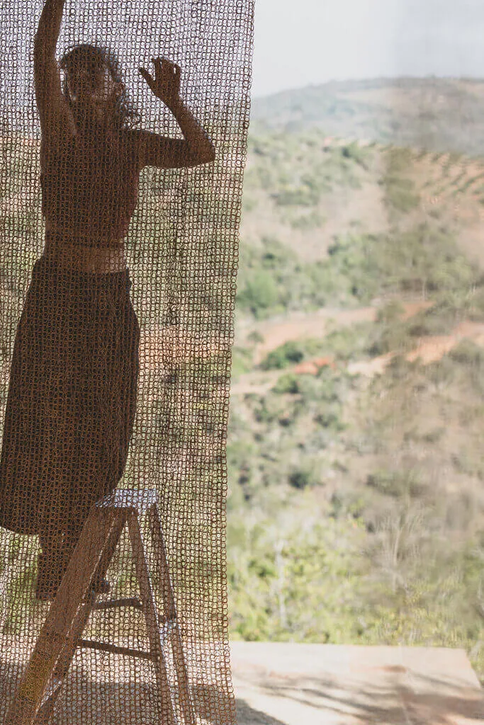 Silhouette of a woman on a ladder hanging Nodomondo sunshade netting with a rural landscape visible in the background.