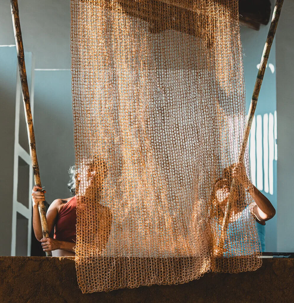 Two people behind a woven net, holding bamboo poles. Sunlight streams through, creating shadows.
