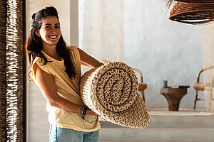 Woman smiling, holding a rolled Nodomondo rug in a bright, stylish interior.
