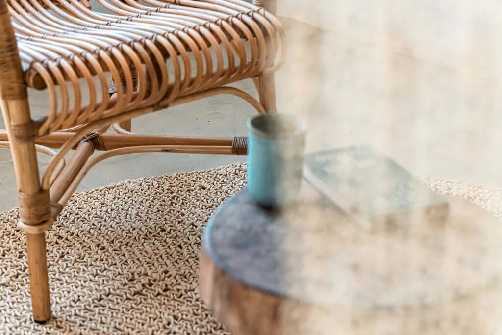 Rattan chair beside a small wooden table with a mug and book on a woven rug.