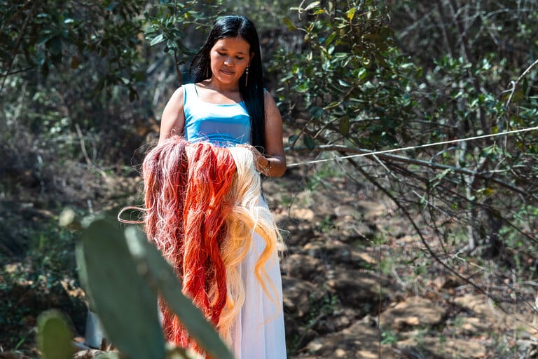 A woman stands in a natural outdoor setting, holding strands of colorful, dyed fibers with green plants and trees around her. VOLVERACASA.