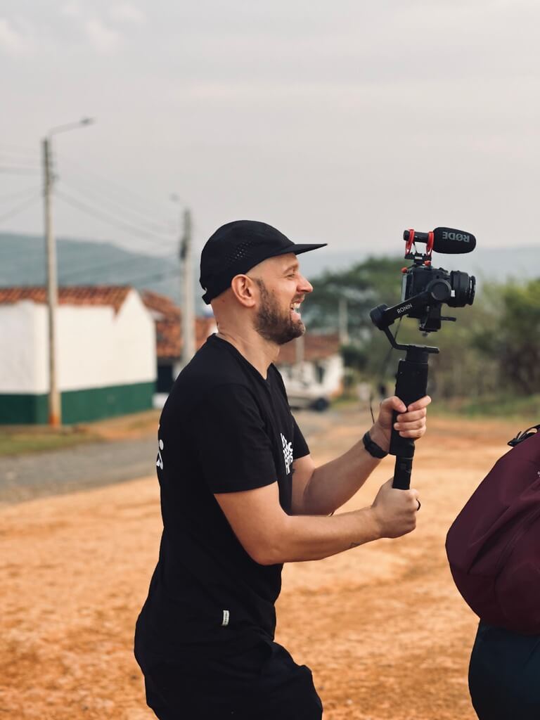A bearded man wearing a black cap and black shirt holds a camera mounted on a stabilizer while filming outdoors on a dirt path, capturing the essence of embodic films.