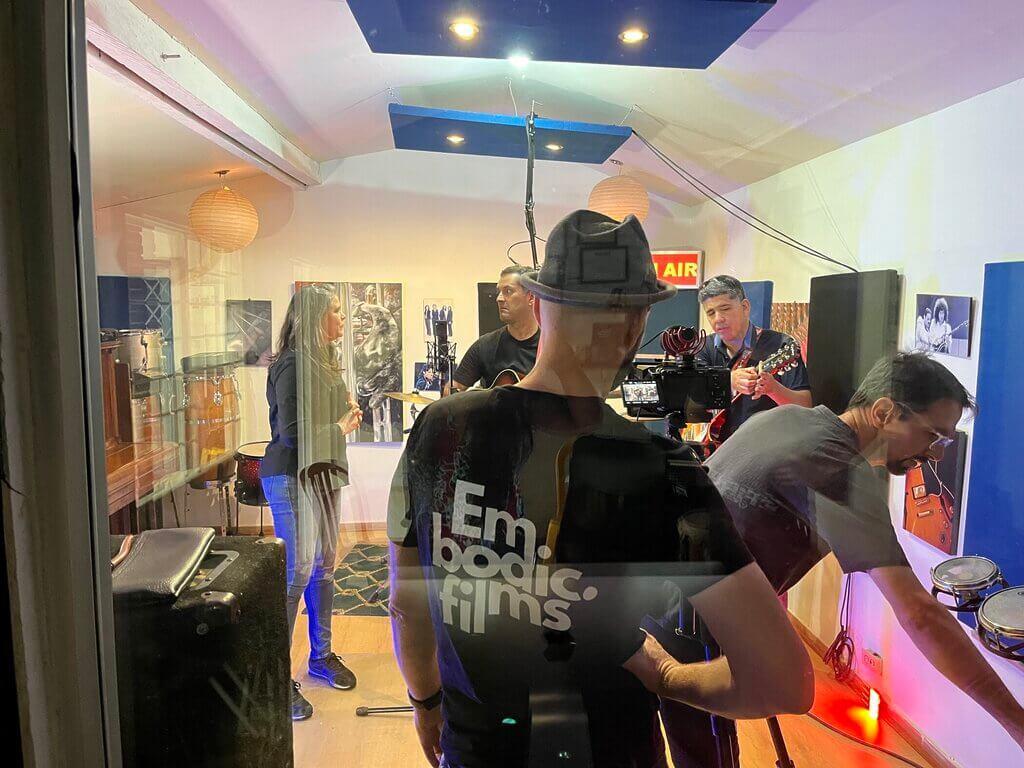 Band recording in a studio, seen through glass. "Embodica films" on a crew member's shirt.