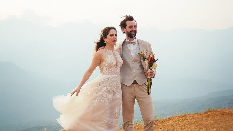 Bride and groom in wedding attire pose outdoors against a mountain backdrop.