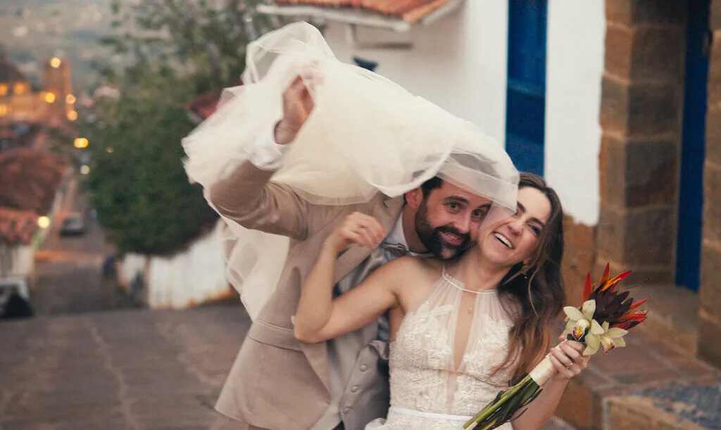 Mariana & Alex wedding photo: Bride and groom playfully under veil with bouquet in a charming town.