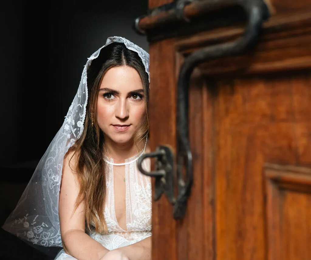 Bride with lace veil and dress peeks from behind a wooden door. Weddings