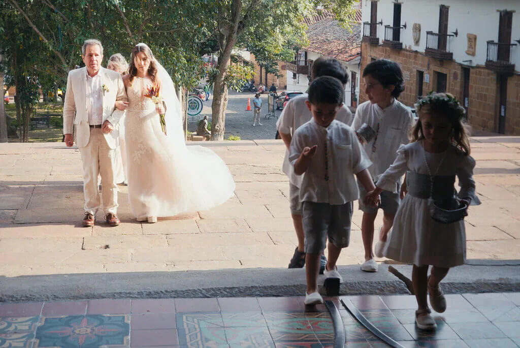 A bride and groom walk up steps towards a building with several children in white attire ahead of them. The scene is outdoors with trees and a tiled floor visible.