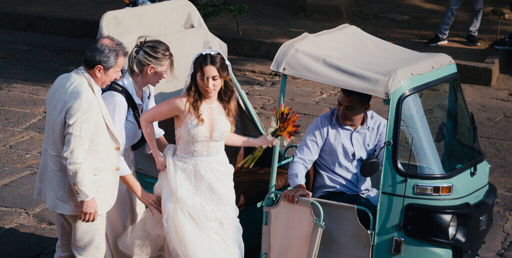 A bride in a white dress steps out of a tuk-tuk, assisted by two men and followed by another woman, holding a bouquet of orange and yellow flowers.