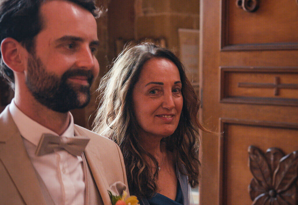 A bearded man in a suit and bow tie stands beside a woman with long brown hair, smiling, in an indoor setting with wooden elements visible in the background.