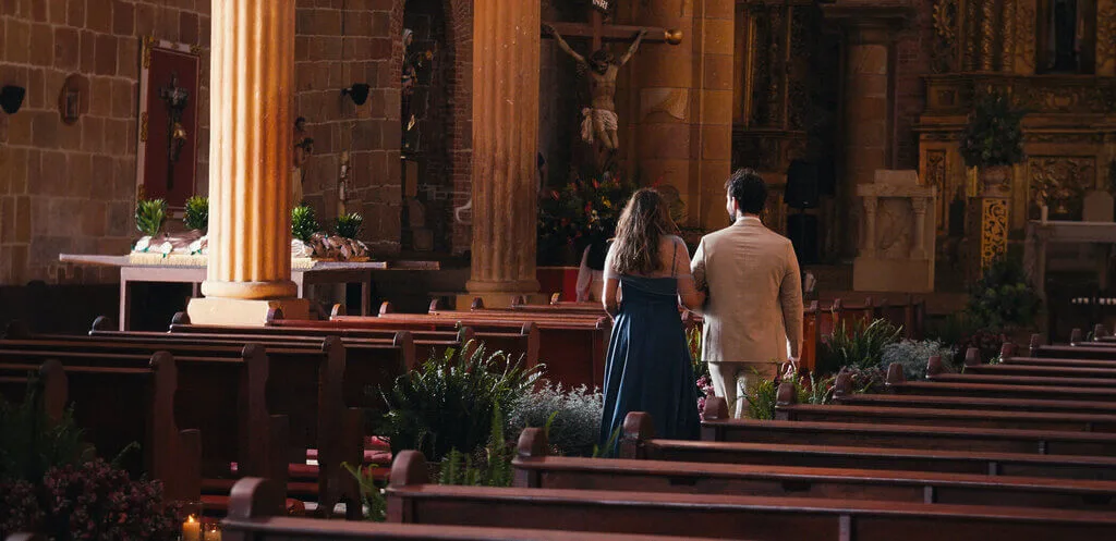 A woman in a navy dress and a man in a white suit walk down the aisle of a dimly lit church with wooden pews and decorations.