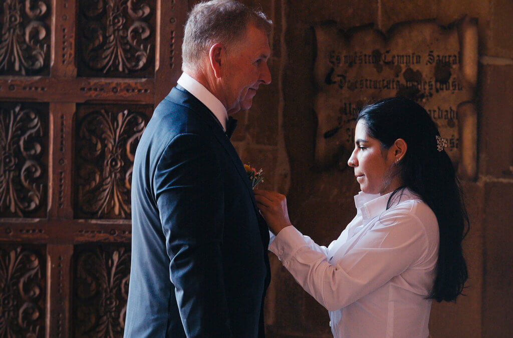A person in a white shirt is pinning a boutonniere onto a person in a suit jacket, standing in a room with ornate wooden doors and a stone wall with a plaque.