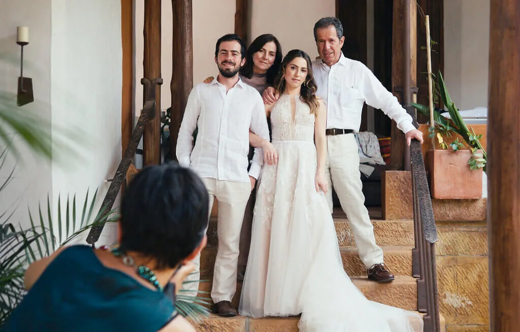 A woman in a white dress poses with three people on wooden steps while another person takes a photo. The setting appears to be indoors with plants in the foreground.