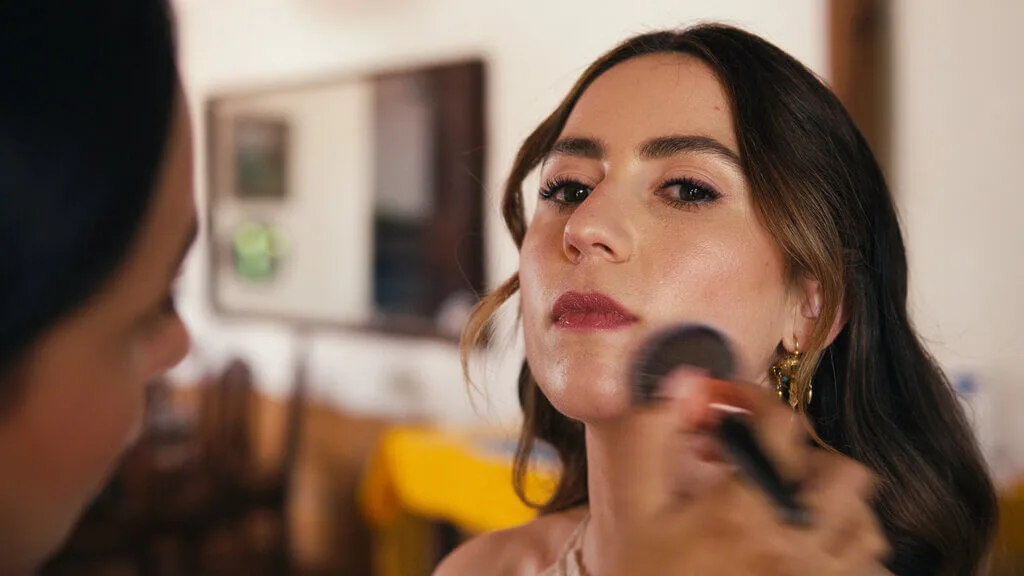 A woman with long brown hair is having makeup applied to her face with a brush by another person. She is looking slightly upwards.