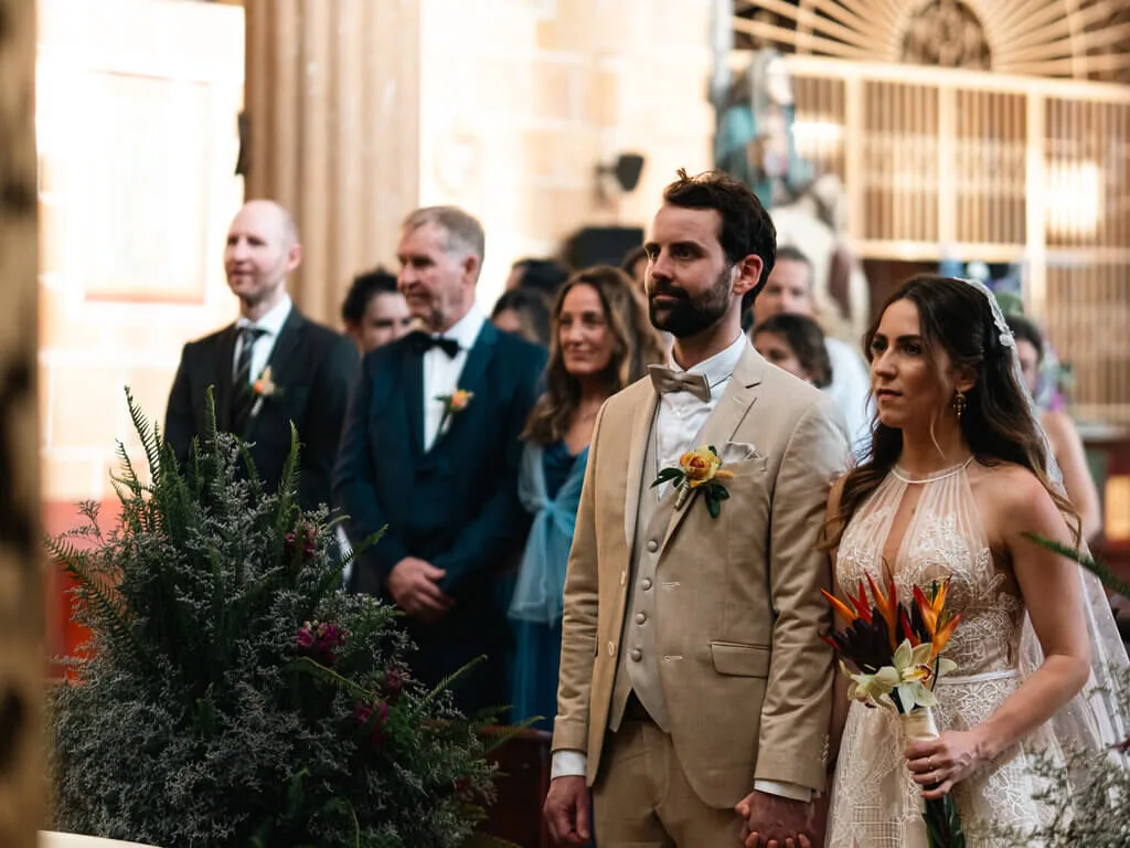 Mariana and Alex at their wedding ceremony, holding hands, with guests in the background.