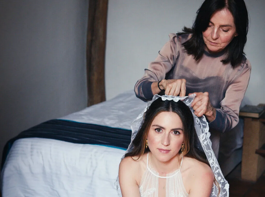 A bride sits in front of a bed while an older woman places a veil on her head.