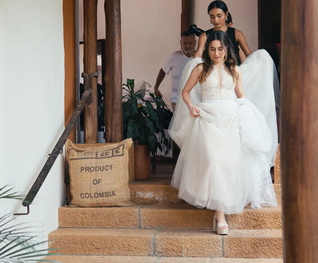 A bride in a white dress descends a staircase with the help of two attendants. A burlap sack labeled "Product of Colombia" is placed beside the steps.