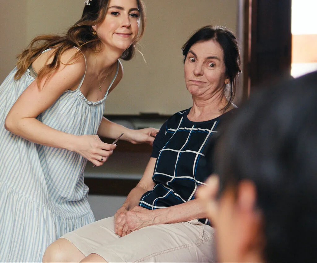 Two women indoors: one with long hair in a striped dress smiles while the other, seated with hands clasped, has short hair and wears a checkered top, looking at the camera with an expressive face.