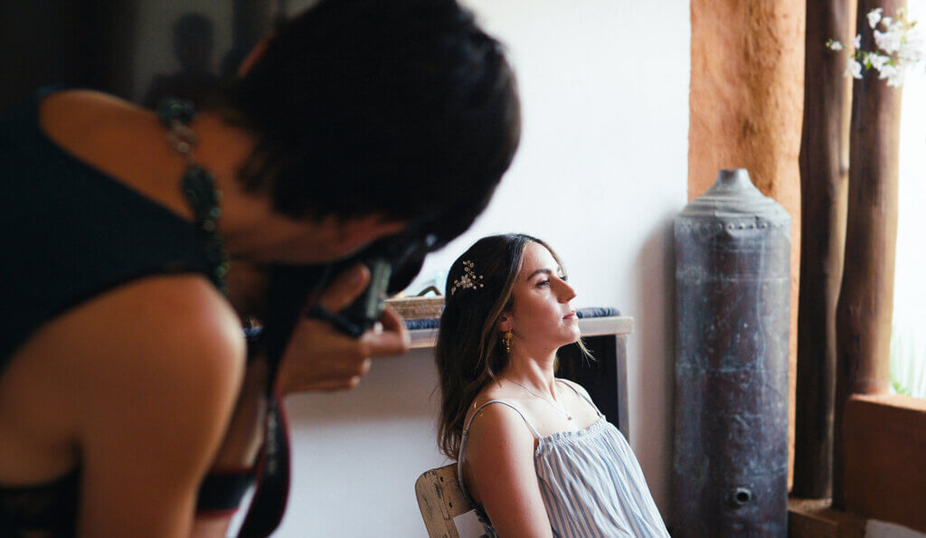 A woman sits on a chair, posing for a photographer who is focusing the camera on her in an indoor setting.
