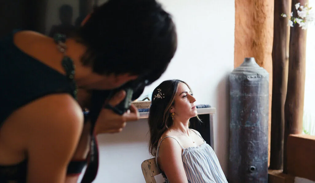 A woman sits on a chair, posing for a photographer who is focusing the camera on her in an indoor setting.