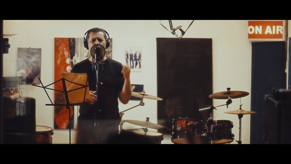 Man singing into a microphone in a recording studio with drums and an "ON AIR" sign.