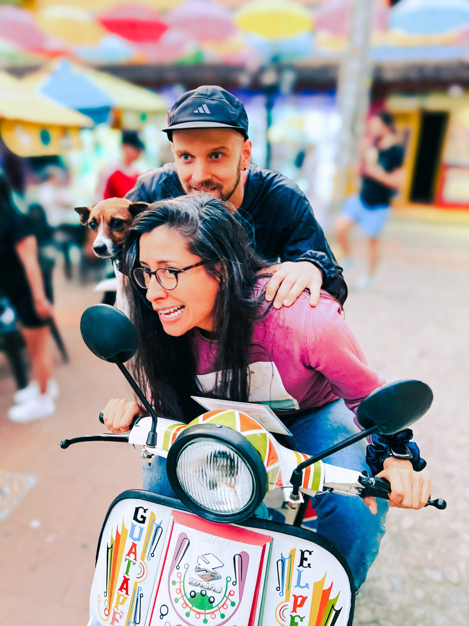 Two people and a dog are riding a decorated scooter. The woman, wearing glasses and a pink shirt, sits beside the man in a cap and jacket. The dog is perched comfortably between them. In the background, colorful umbrellas add to the festive scene, creating an atmosphere about joy and companionship.