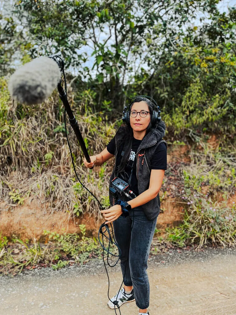A person holding a boom microphone and wearing headphones stands on a gravel path, surrounded by greenery. They appear to be monitoring audio equipment.