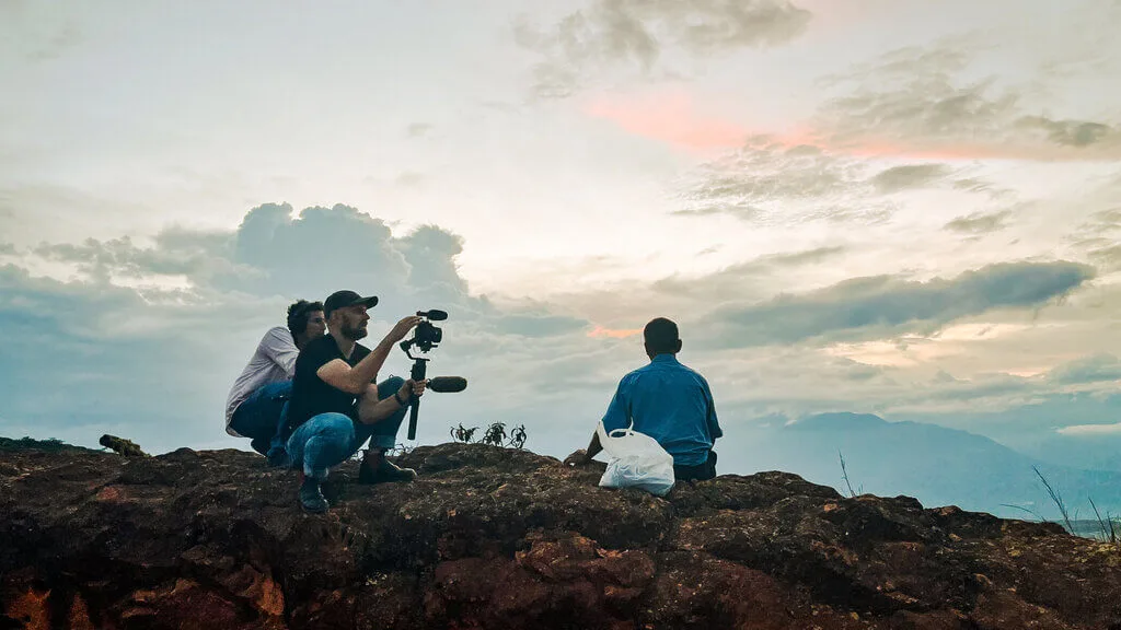 Two men with camera equipment film a person sitting on a rocky hill under a cloudy sky.