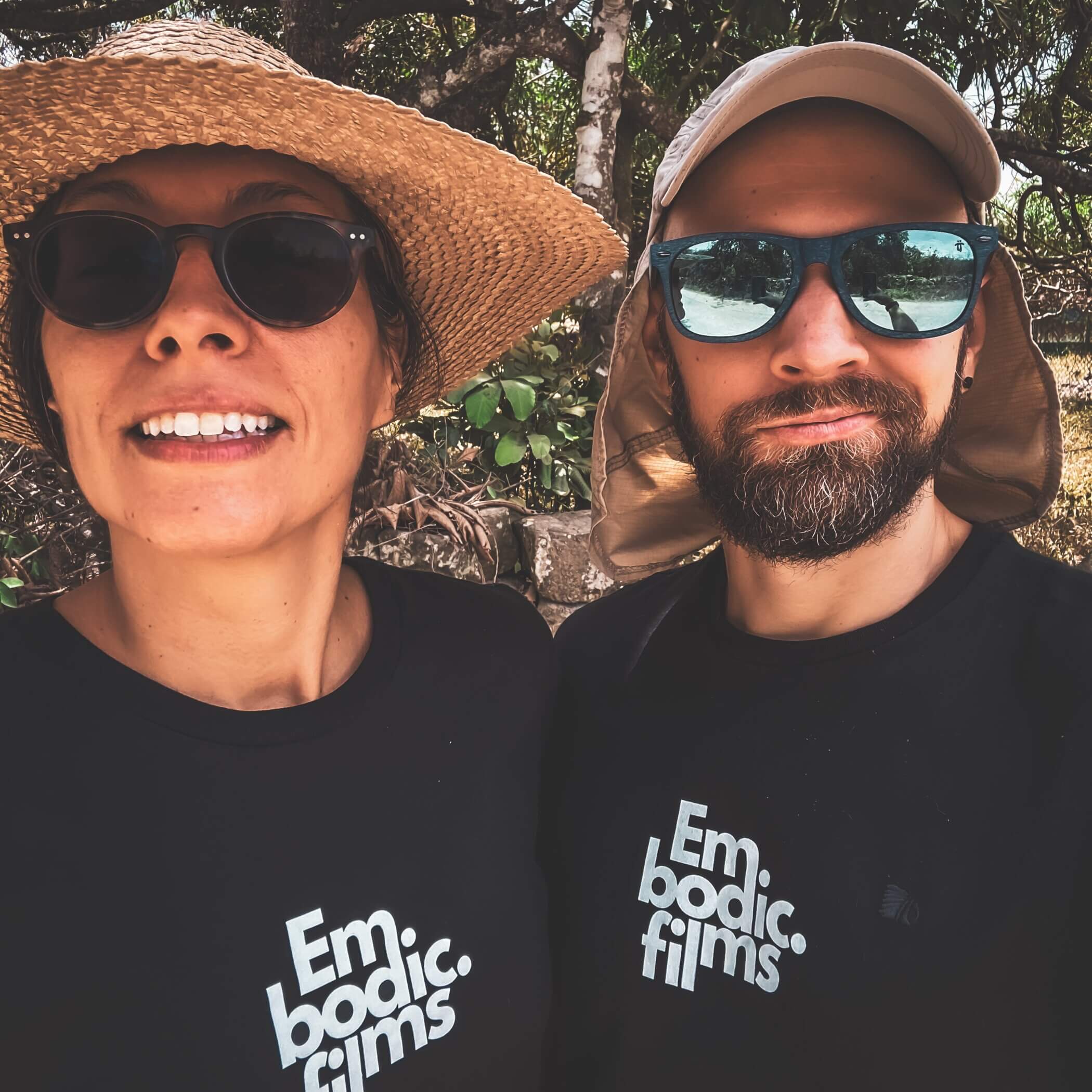 Two people, a woman and a man, pose outdoors wearing sunglasses and black shirts with the text "Embodic Films". The woman has a straw hat, and the man wears a cap with a neck cover.