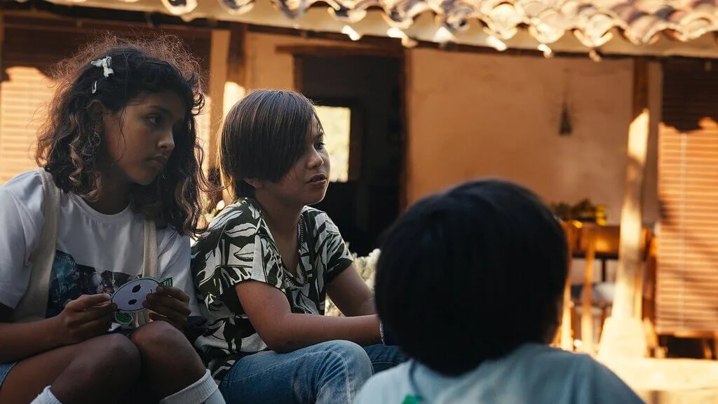 Three children sit closely together outside a rustic building. Two face forward, one holding a small object, while the third’s back faces the camera. Warm sunlight and an Embodic Films touch illuminate this heartfelt scene.