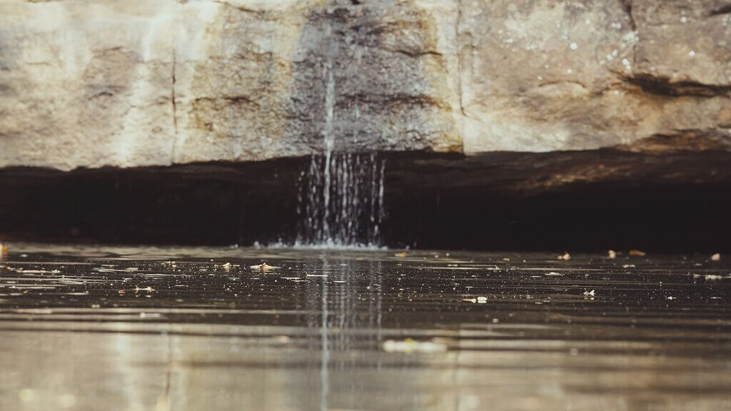 A small stream of water gently falls from a rocky ledge above into a calm, shallow pool below, creating ripples on the reflective water surface—a tranquil moment captured by Embodic Films.