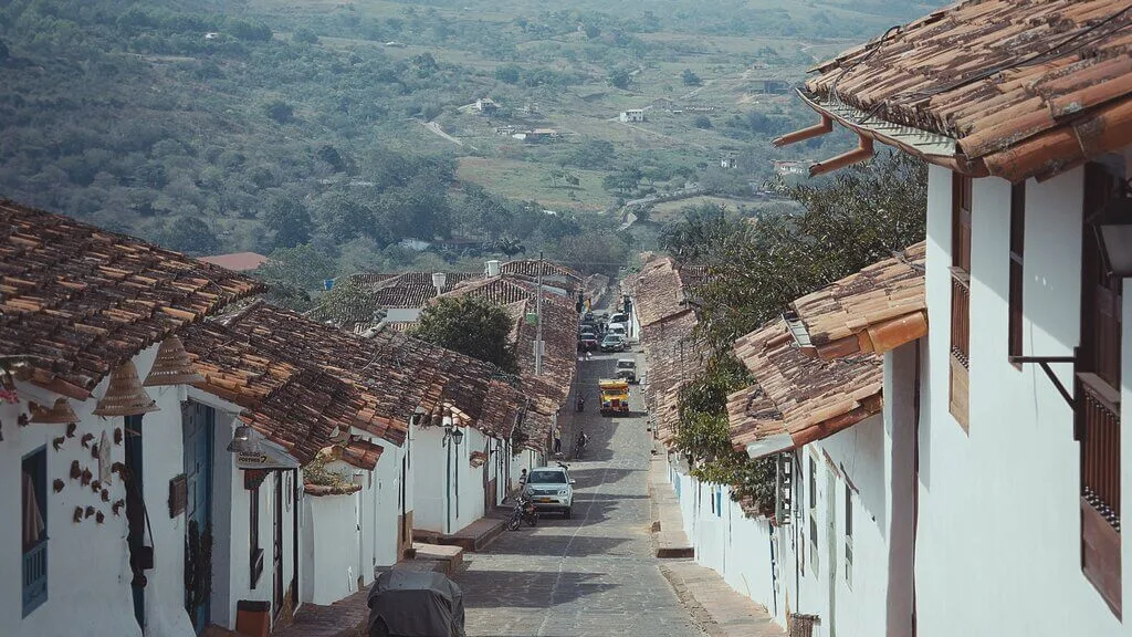 A narrow cobbled street lined with white buildings and terracotta roofs descends a hill in Barichara, Colombia. Cars are parked along the street, and greenery covers the hilly background.