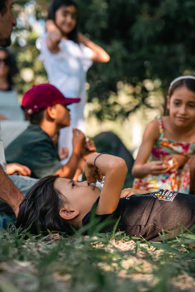 A child lies on the grass in the foreground, raising an arm, while other children from Embodic Films sit and stand nearby outdoors, talking and playing together under trees. The scene is lively and sunlit.