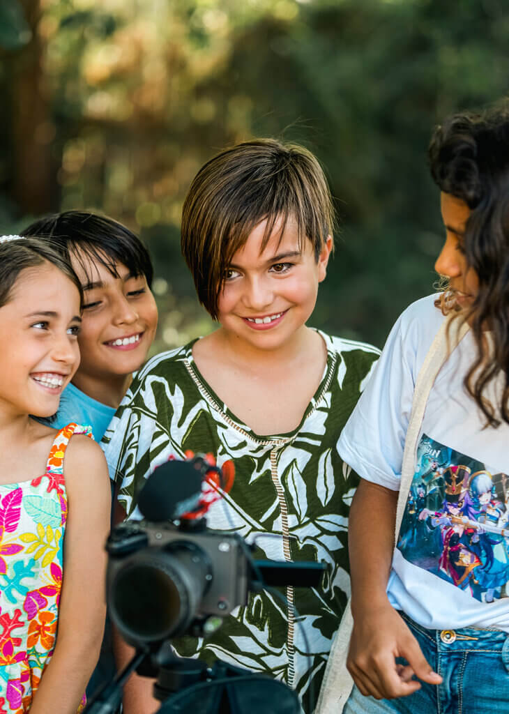 Four smiling children stand outdoors around a camera on a tripod, capturing a joyful moment for Embodic Films. Surrounded by greenery, one child looks directly at the camera while the others interact and laugh together.