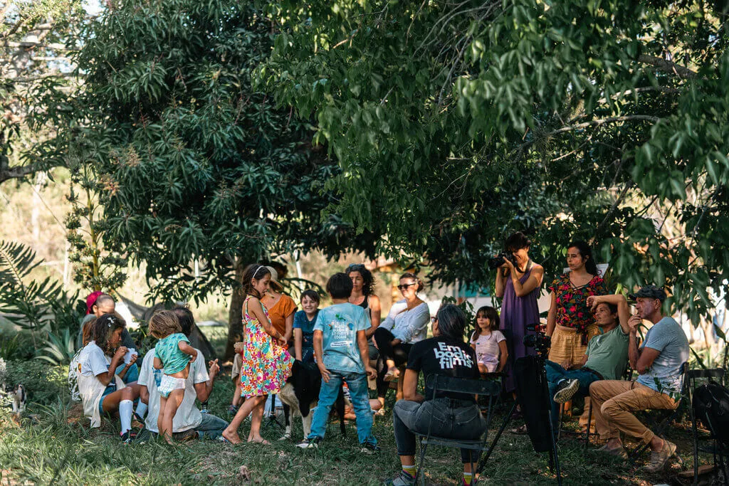 A group of people, including children and adults, are gathered outdoors under the shade of trees, engaged in conversation and activities. Some are seated, while others stand or interact with children.