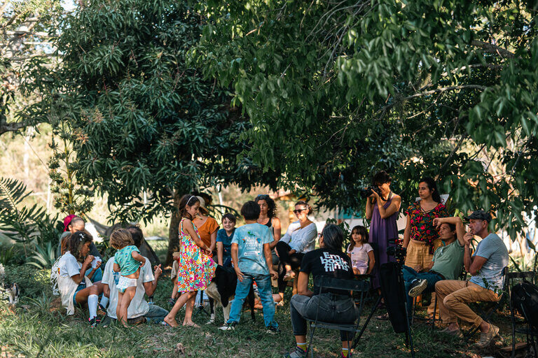 A group of people, including children and adults, are gathered outdoors under the shade of trees, engaged in conversation and activities. Some are seated, while others stand or interact with children.