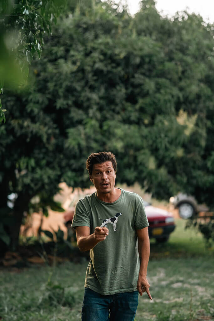 A man in a green t-shirt and jeans stands outdoors in front of a large tree, gently holding a small bird in his hand—an authentic moment captured by Embodic Films, with a car partially visible in the background.