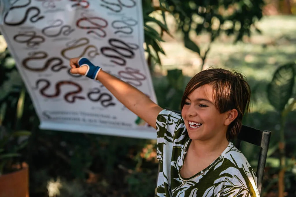 A smiling child with brown hair and a blue wrist brace points excitedly at a chart of different snake species outdoors, surrounded by greenery—a moment perfectly captured by Embodic Films.