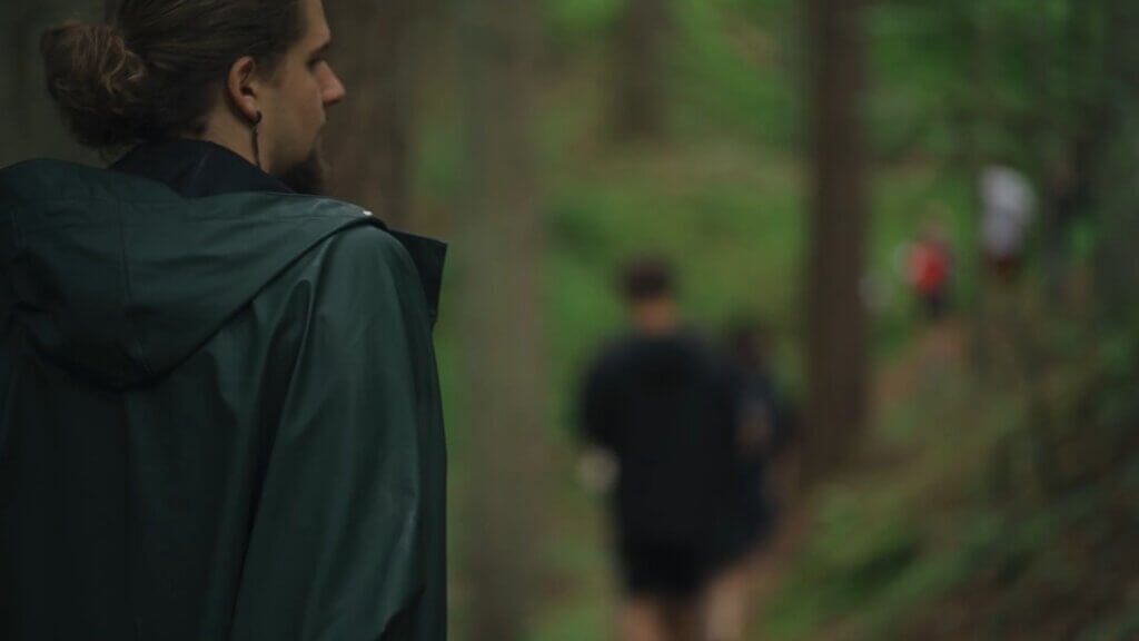 Person in a green raincoat watching others on a forest trail. Shinrinyoku.lt