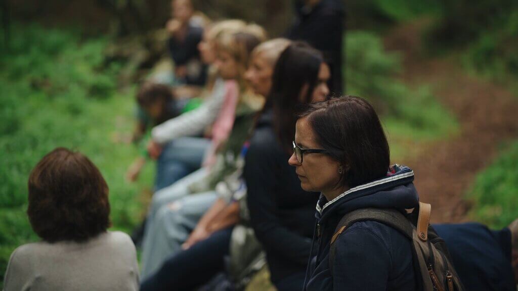 People sitting outdoors, possibly experiencing Shinrinyoku, with a woman in glasses in the foreground.