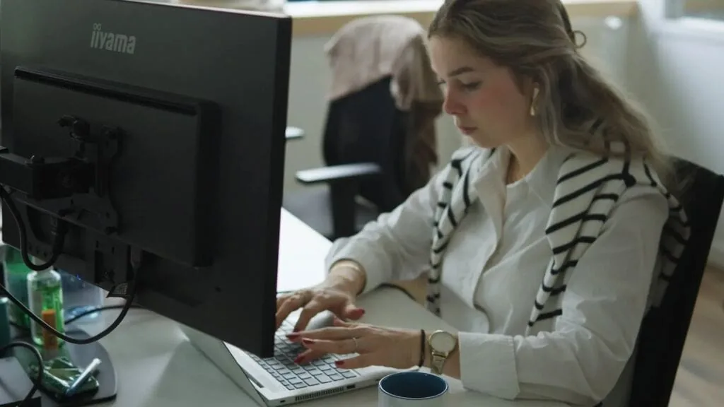 Woman working on a laptop at a desk with a monitor.