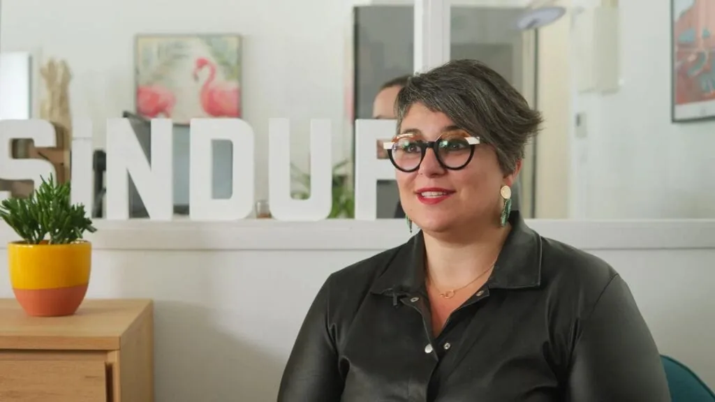 Woman with glasses in front of Sindup logo. Office setting.