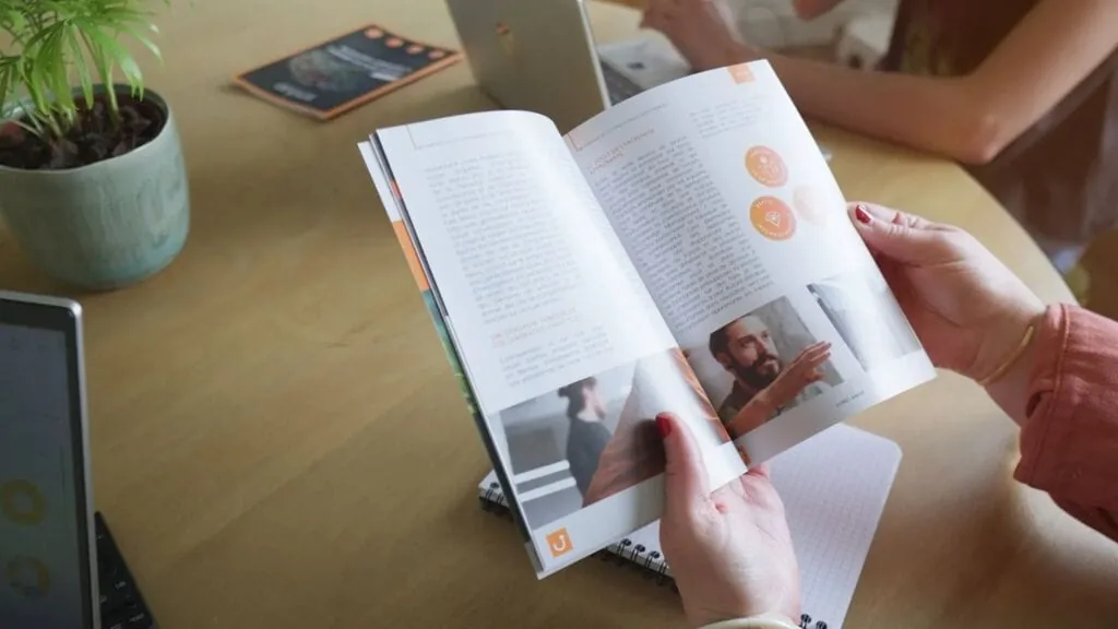 Hands holding a Sindup brochure open on a desk with a laptop and plant.