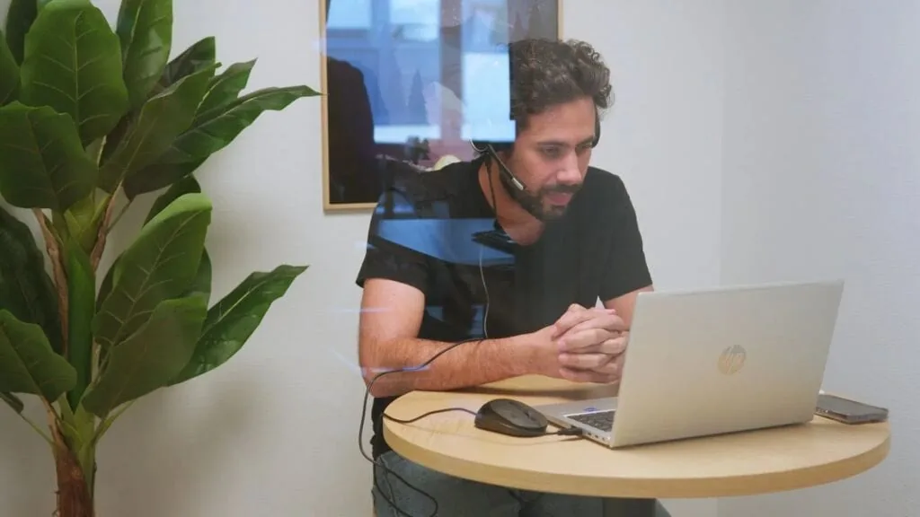 Man with headset working on laptop at desk with plant.